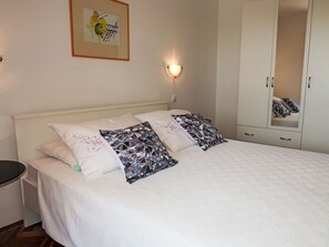 Un letto rifatto con cura, lenzuola bianche e cuscini decorati, un comodino, una lampada a parete e un armadio con specchio.