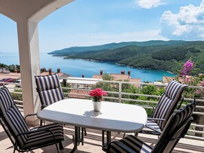 Une terrasse avec une table et des chaises, donnant sur un paysage côtier avec des collines et une étendue d’eau.