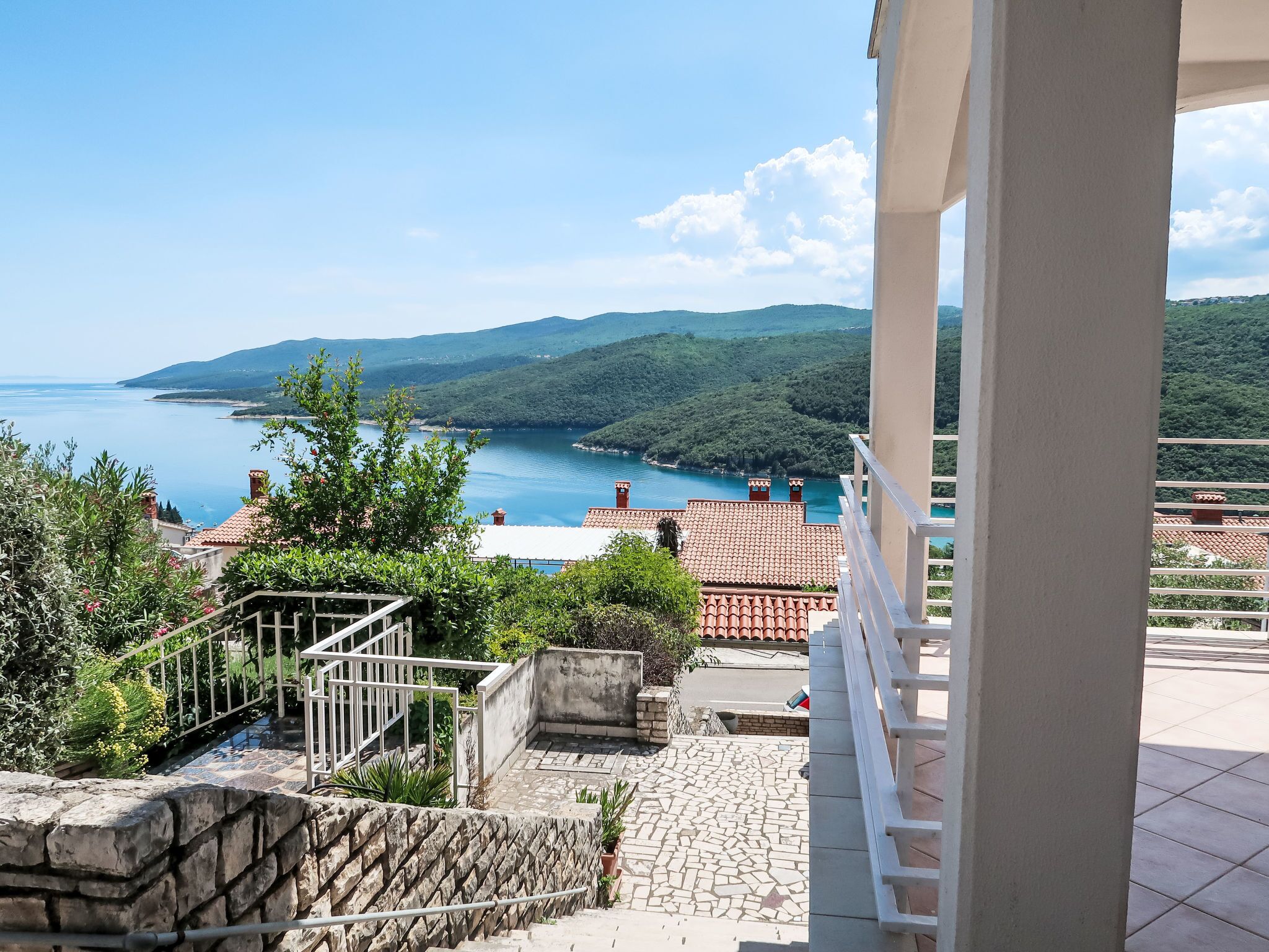 A balcony with a stone wall and metal railing overlooking a coastal landscape with a clear view of the sea and hills.