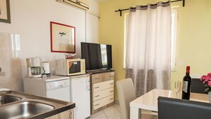 Een compacte keuken met een magnetron, koffiezetapparaat en een televisie op een houten kast. Een zithoek met een tafel en stoelen is zichtbaar, evenals een raam met gordijnen.