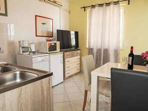 Une cuisine compacte équipée d’un four à micro-ondes, d’une machine à café et d’un téléviseur posé sur un meuble en bois. On aperçoit un coin repas avec une table et des chaises, ainsi qu’une fenêtre avec des rideaux.
