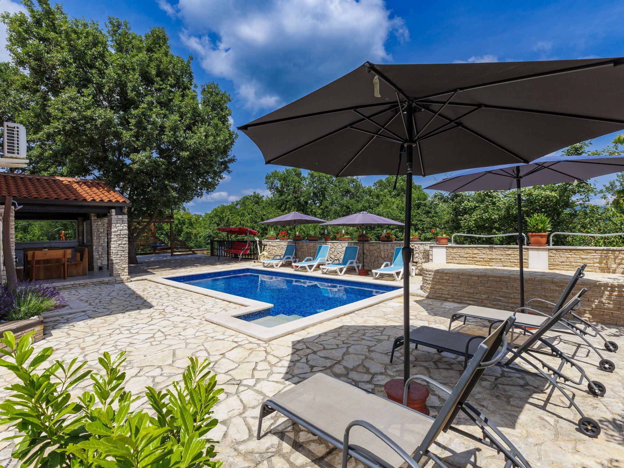Piscina con sillas de descanso y sombrillas, rodeada de pavimento de piedra y vegetación.