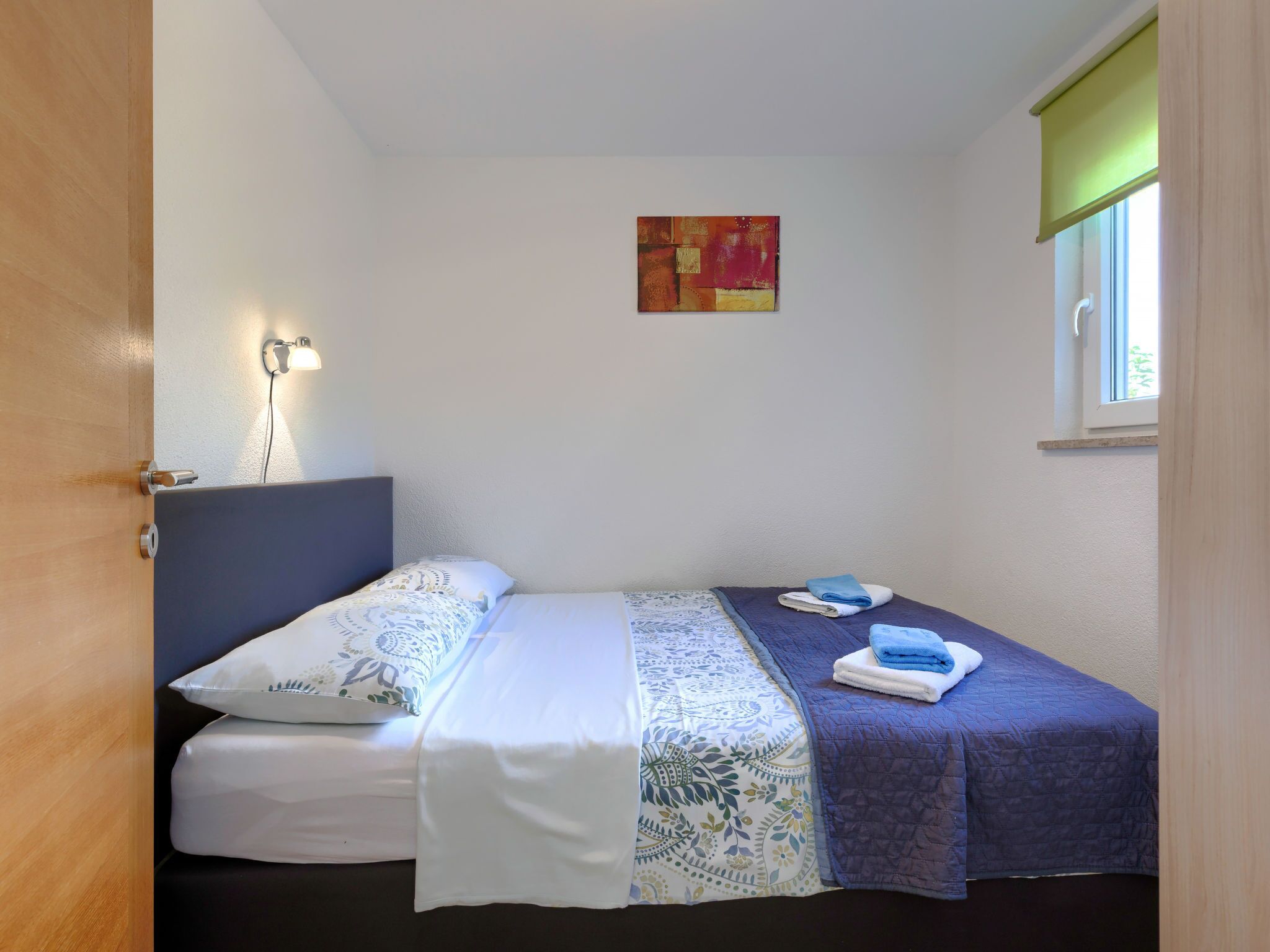 Ein Einzelbett mit gemustertem Bettbezug, eine Nachttischlampe, ein Fenster mit grüner Jalousie und ein kleines Bild an der Wand.