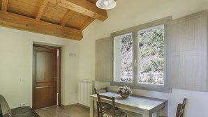 Een kamer met een houten plafond, een eettafel met stoelen, een raam met uitzicht op groen, en een deur.