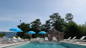 A swimming pool with lounge chairs and blue umbrellas, surrounded by trees and a stone structure. - Vacation home Diana by Interhome (Labin)
