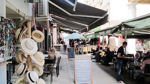 Un caffè all'aperto con tavoli e sedie sotto ombrelloni, un espositore per cappelli e una lavagna con il menu.