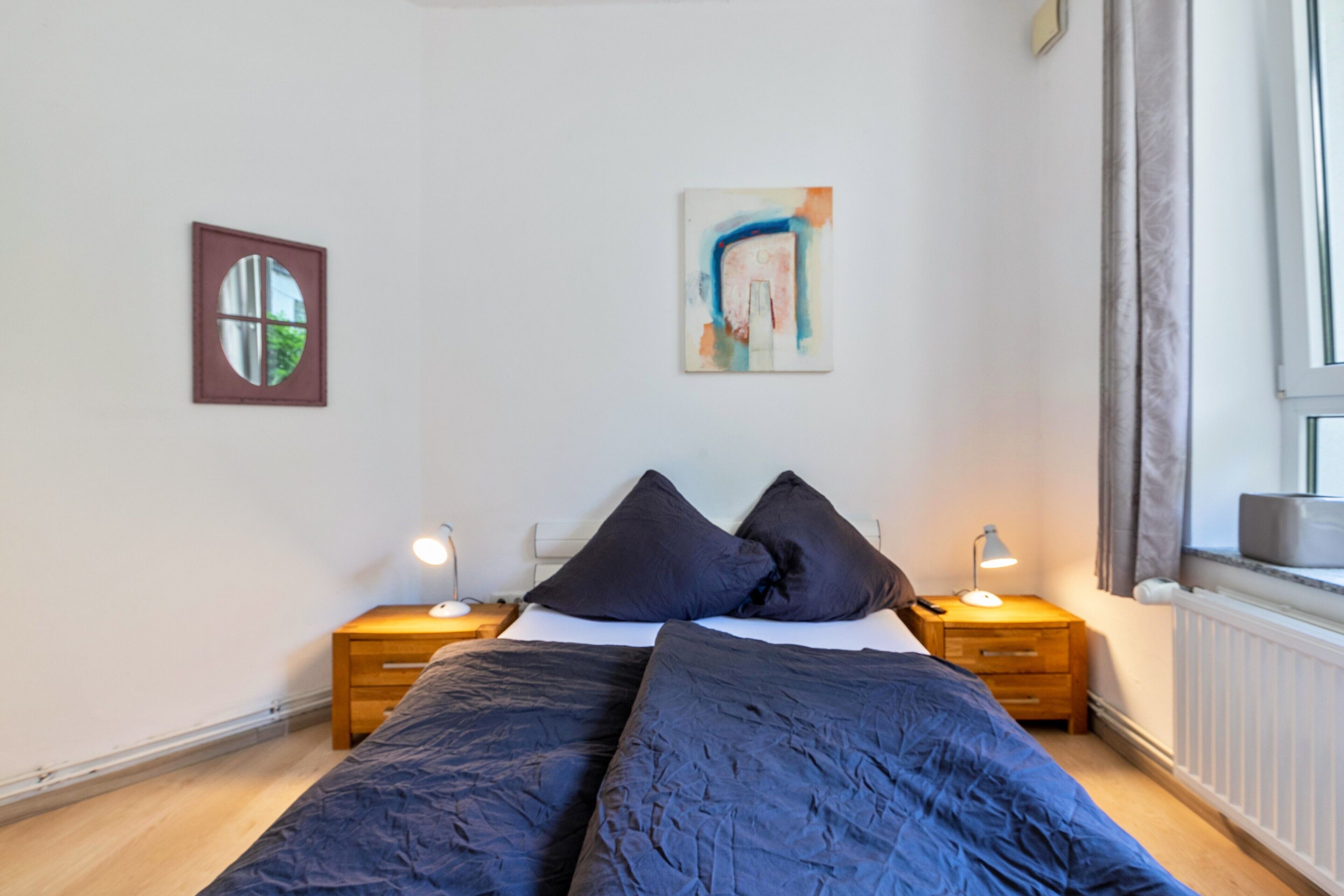 Une chambre à coucher avec un lit, deux tables de chevet, une fenêtre et des œuvres d’art accrochées au mur.