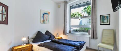 Een slaapkamer met een bed, nachtkastje, lamp, raam, stoel en kunstwerk.