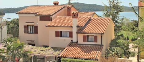 A multi-story building with a tiled roof, surrounded by greenery and a body of water in the background.