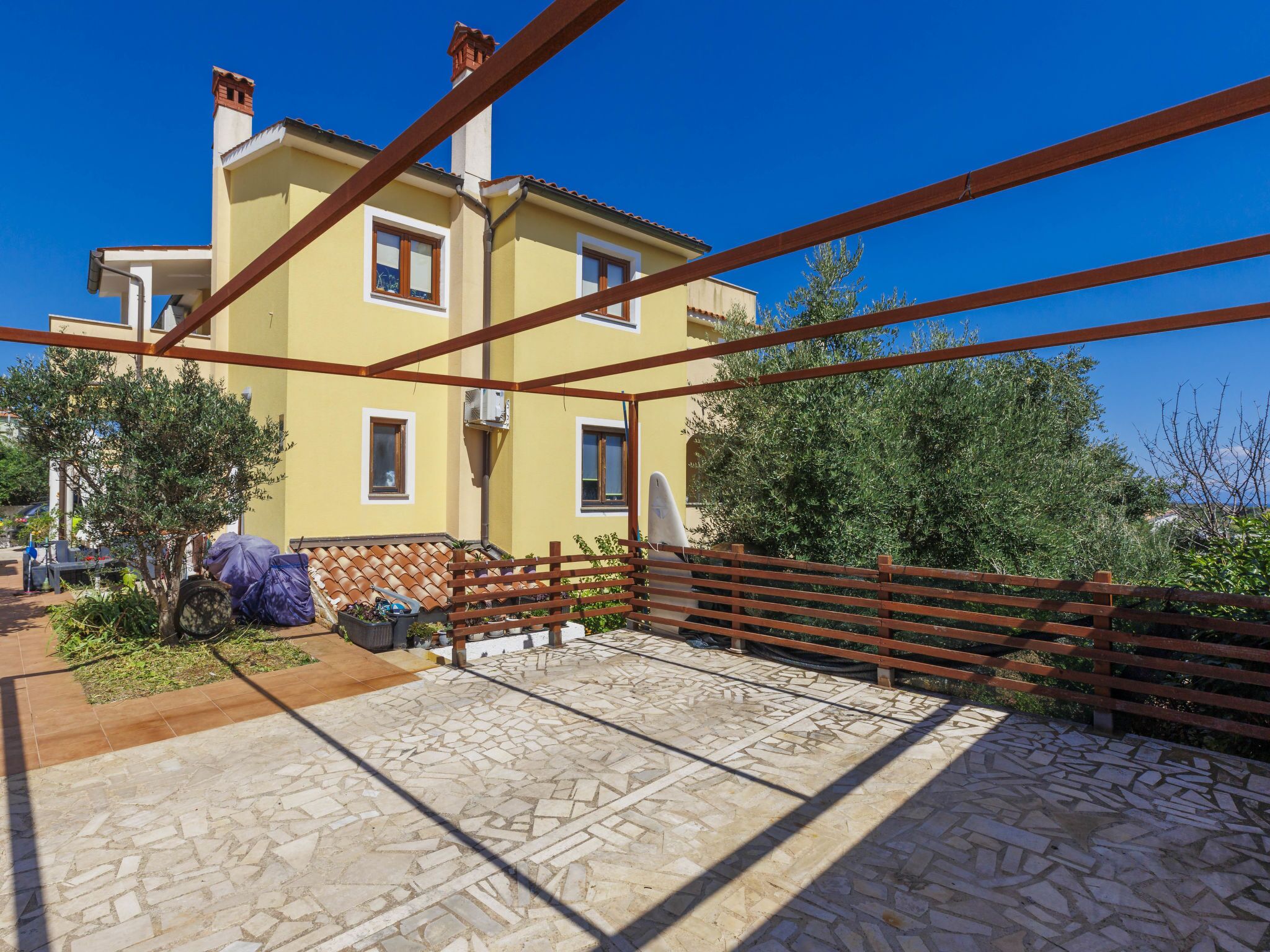 Une vaste terrasse dotée d’un sol carrelé, d’une clôture en bois et d’un bâtiment jaune avec de nombreuses fenêtres.