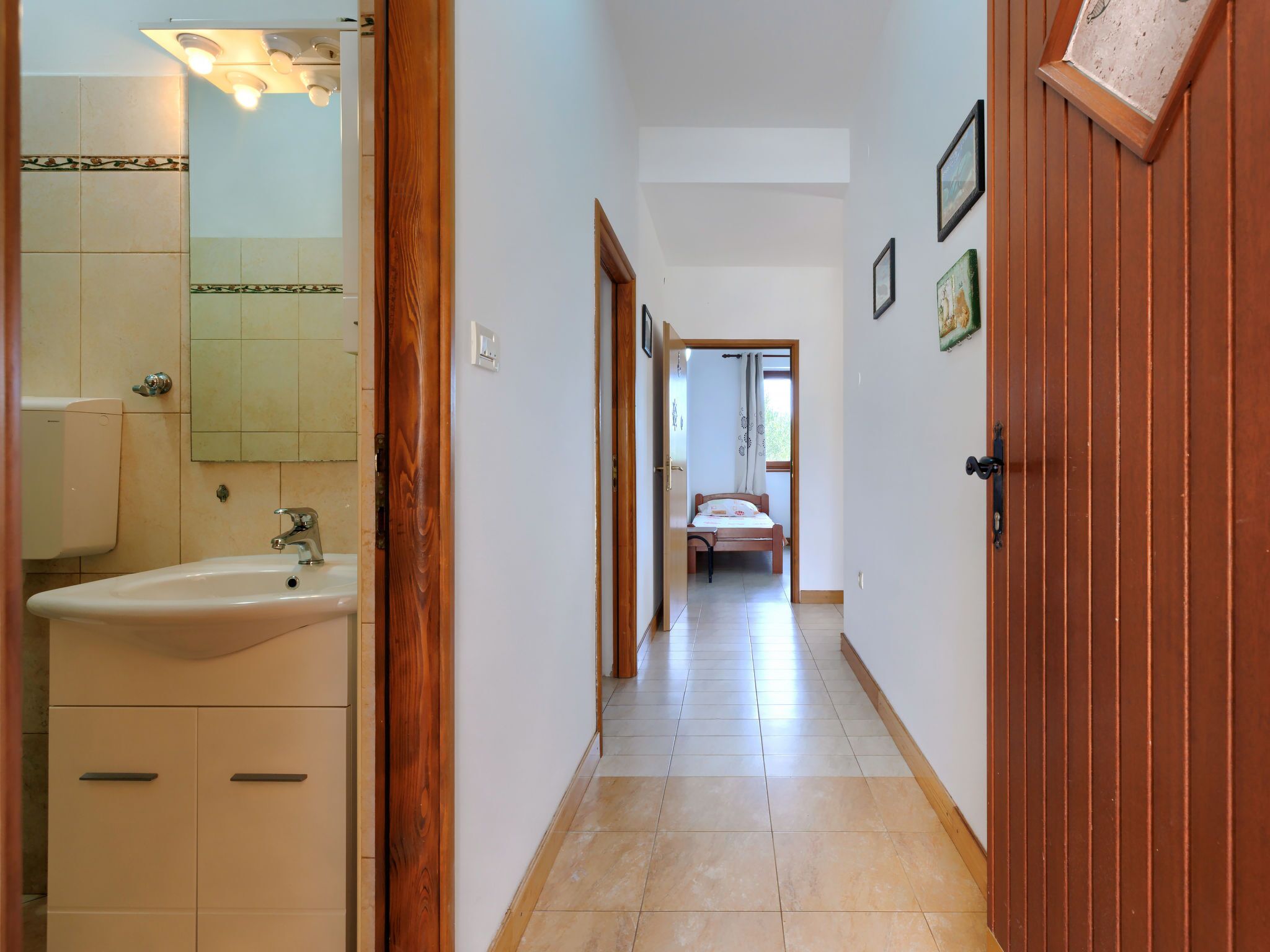 A narrow hallway with a bathroom on the left, a bed at the end, and a wooden door on the right.