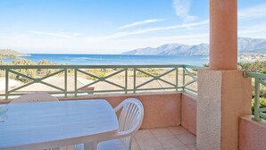 Ein Balkon mit Tisch und Stühlen, der auf eine Küstenlandschaft mit Bergen im Hintergrund blickt.