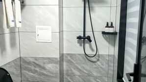 Un baño con azulejos grises y blancos, una ducha con manguera negra y un estante pequeño con artículos de aseo.