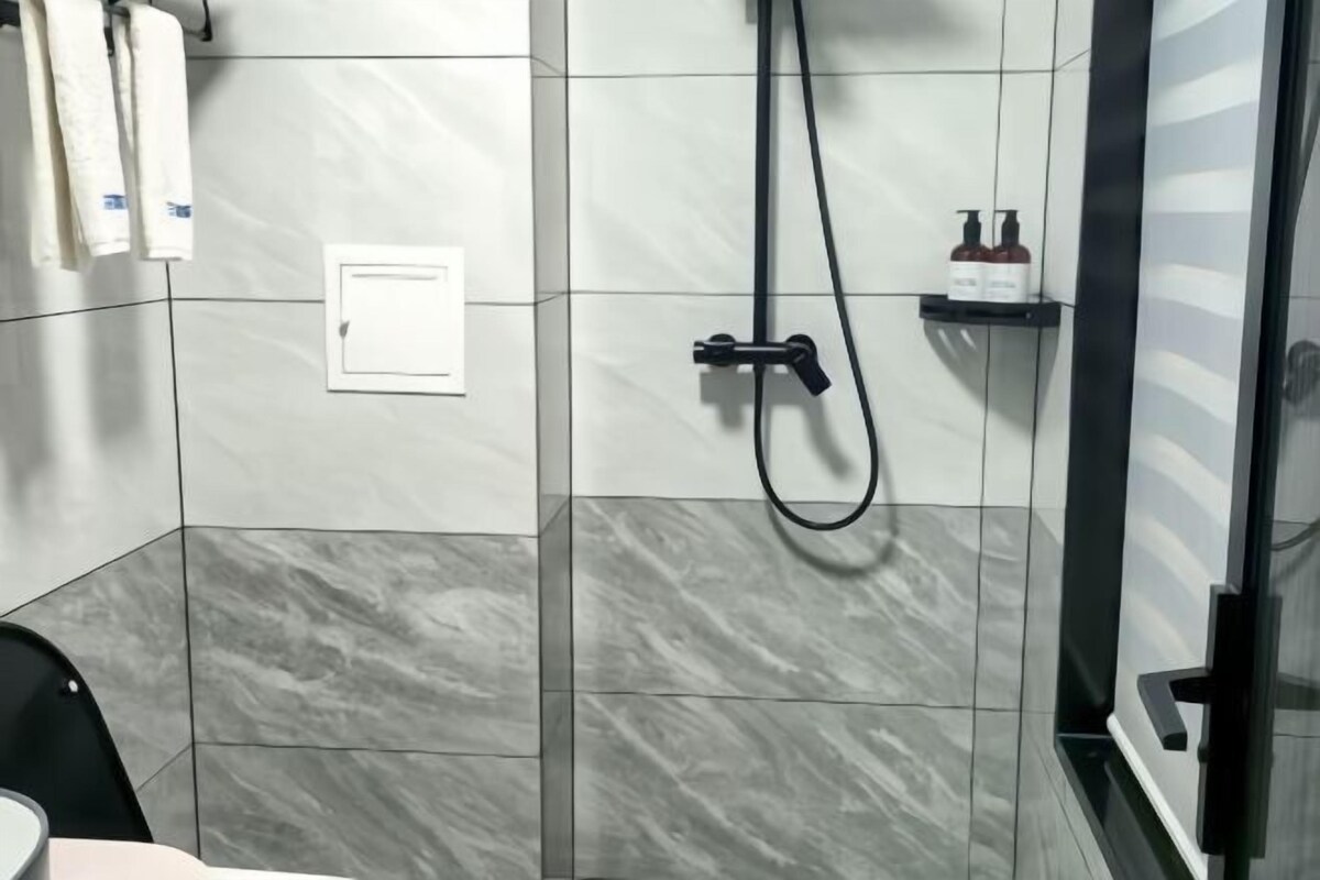 A bathroom with grey and white tiles, a shower with a black hose, and a small shelf with toiletries.