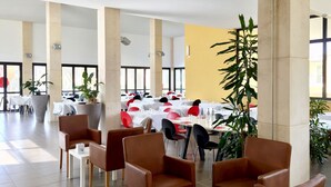 A spacious indoor dining area with high ceilings, large windows, and a yellow accent wall. Tables are set with white tablecloths and red chairs.