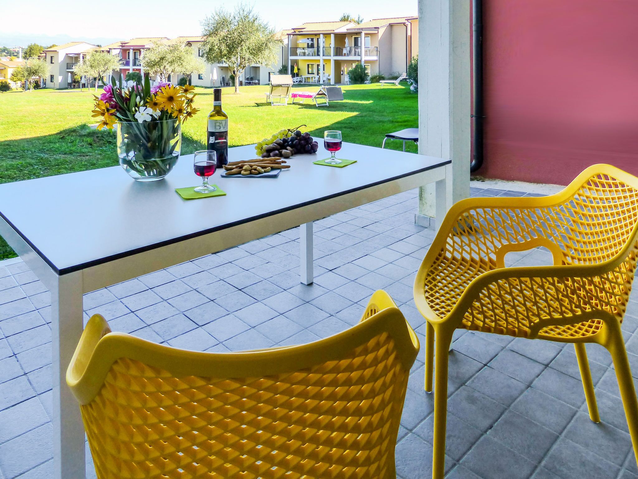 Mesa dispuesta con una botella, copas de vino, uvas y un plato de palitos de pan en un balcón con sillas amarillas y vista a una zona residencial.