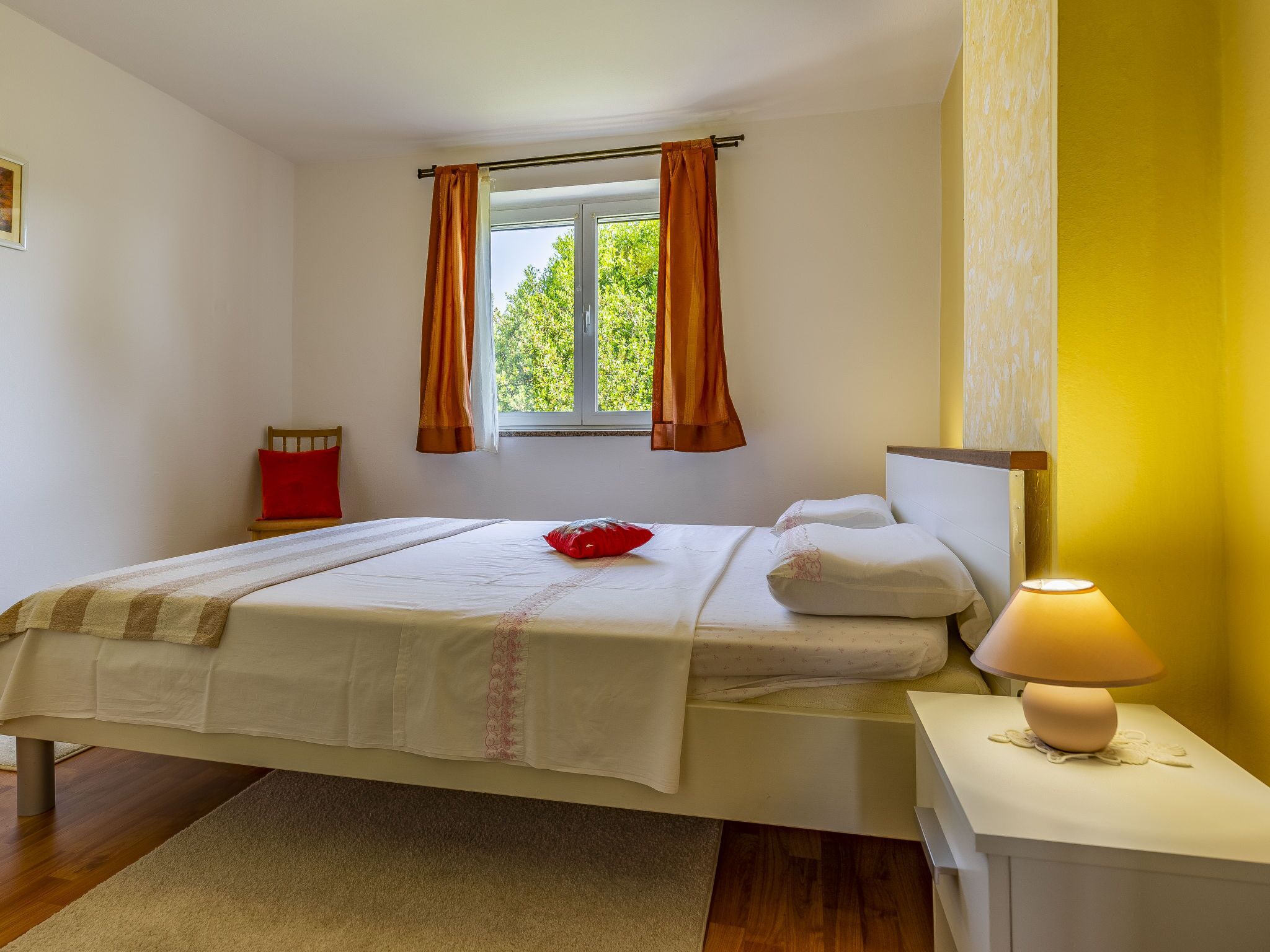 A bedroom with a bed, a nightstand, a window with curtains, and a view of greenery outside.
