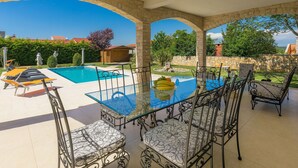 A covered outdoor area with a pool, lounge chairs, and a table with chairs.