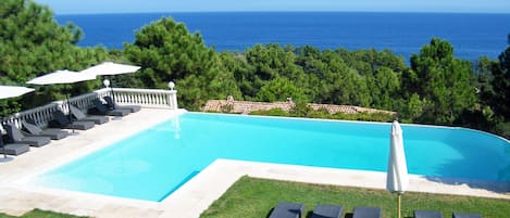 A swimming pool with lounge chairs and umbrellas, surrounded by greenery and overlooking the sea.