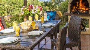 Un espace extérieur aménagé pour les repas, avec vue sur l’océan, un foyer, des meubles en osier et une table dressée avec des assiettes, des verres et du pain.