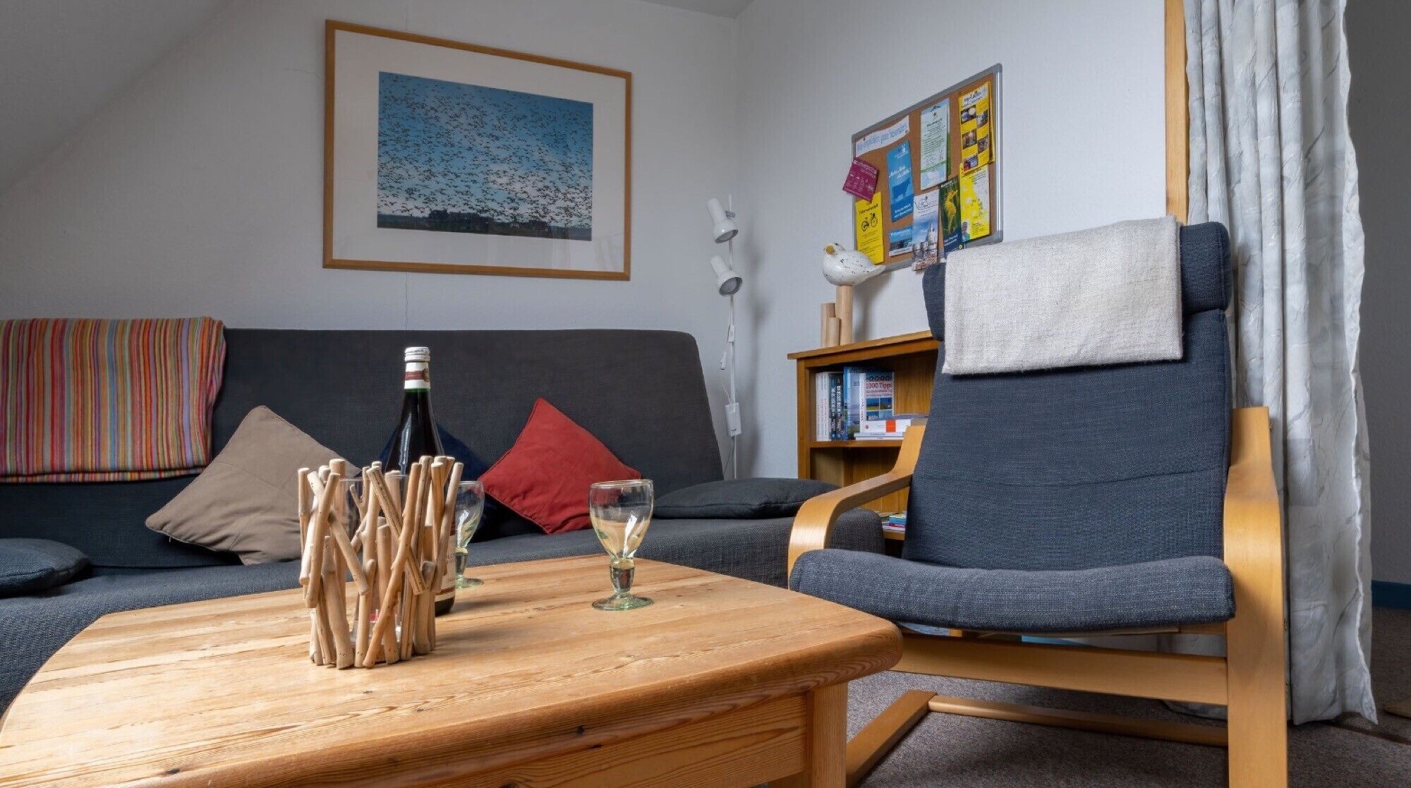 A living room with a sofa, a wooden coffee table with a bottle and glasses, a chair, a bookshelf, and a bulletin board.
