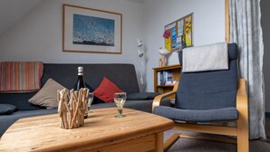 En stue med sofa, sofabord i træ med flaske og glas, stol, reol og opslagstavle.
