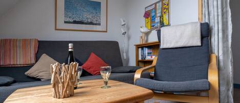 A living room with a sofa, a wooden coffee table with a bottle and glasses, a chair, a bookshelf, and a bulletin board.
