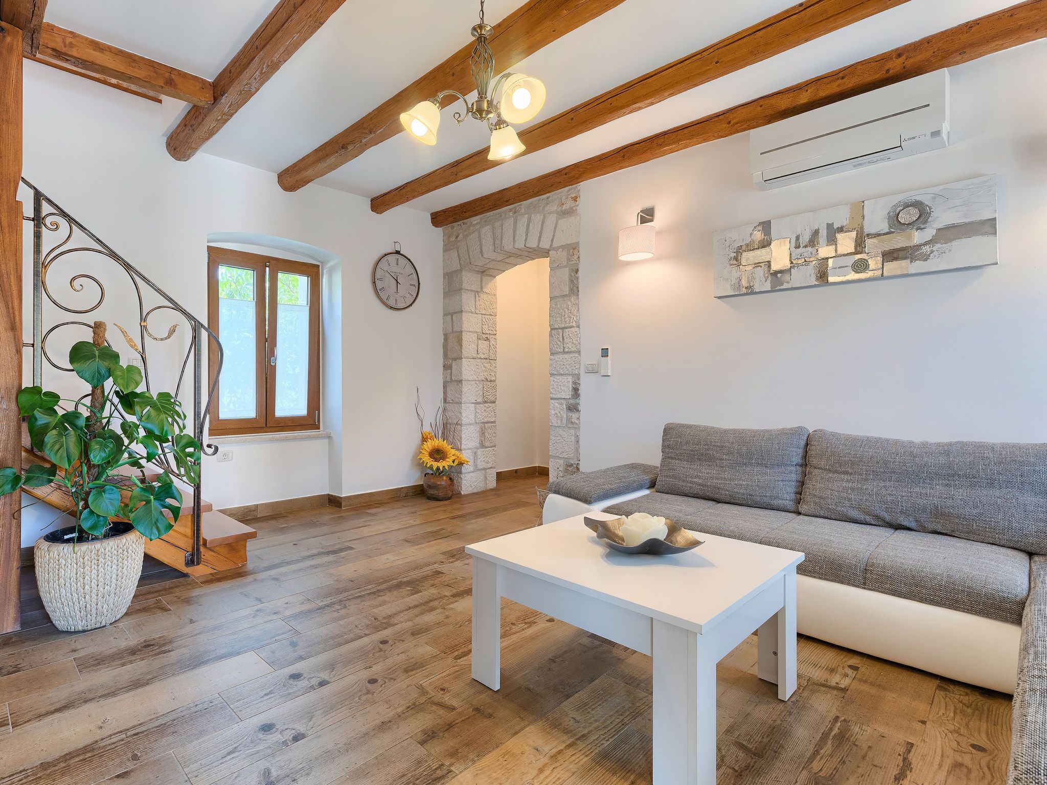 A living room with a white sofa, a white coffee table, wooden flooring, a staircase with a plant, and a stone wall.