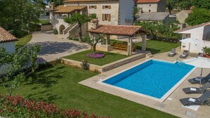 A pool area with a pergola, lounge chairs, and a stone building with a tiled roof.