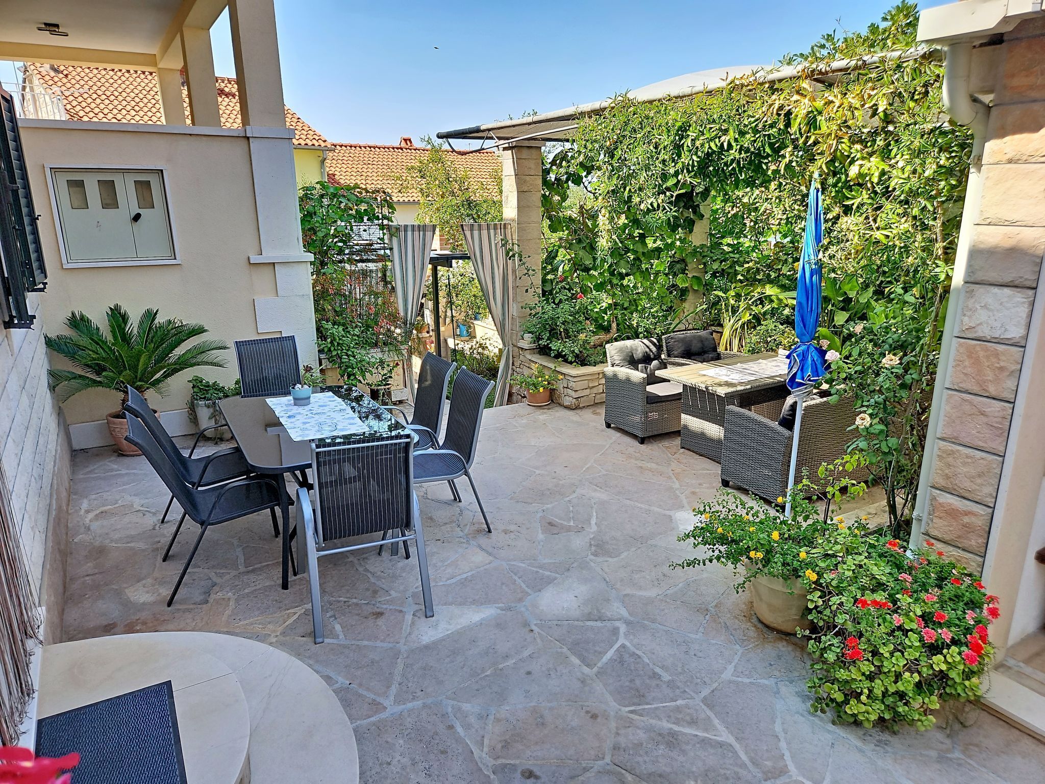 Une terrasse extérieure avec un sol en pierre, une table entourée de chaises, ainsi qu’une variété de plantes et de fleurs en pot.