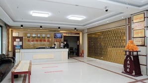 A hotel reception area with a reception desk, a waiting area with a sofa and a table, and a decorative pillar with a plaque. - Wudi Business Hotel (Hunchun)