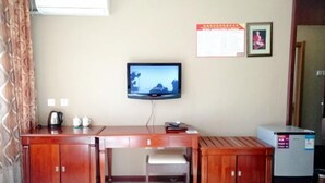 A room with a wooden desk, a television mounted on the wall, a small cabinet, and a refrigerator. - Wudi Business Hotel (Hunchun)
