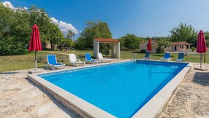 A rectangular swimming pool with clear blue water, surrounded by stone tiles. There are lounge chairs and red umbrellas around the pool. - Danijela by Interhome (Rovinj)