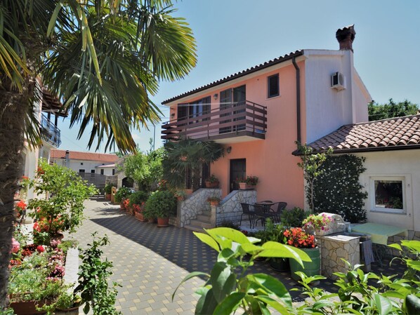 A two-story building with a balcony, surrounded by potted plants and a paved courtyard.
