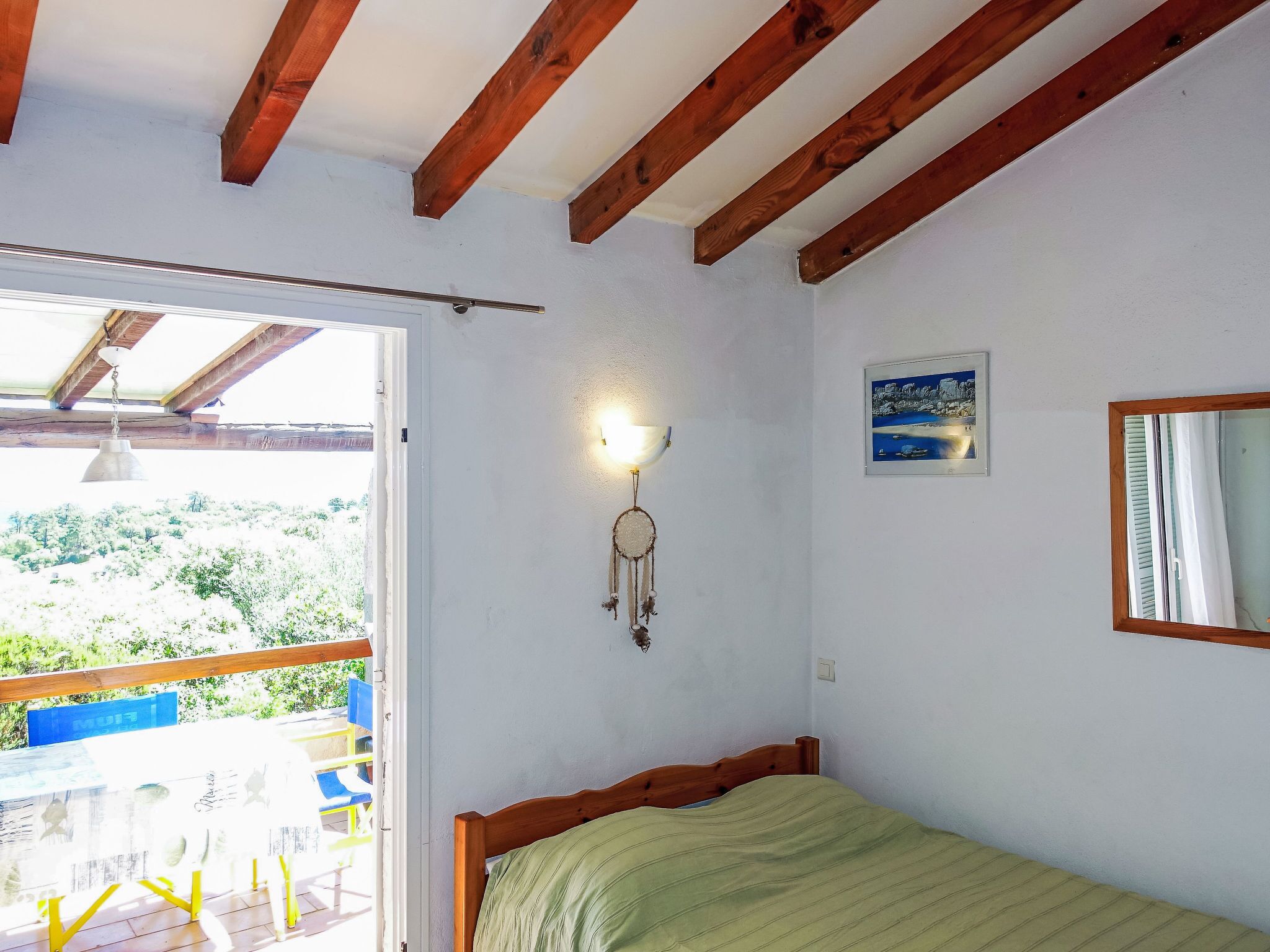 Ein Schlafzimmer mit Holzbalkendecke, einem Bett, einer Wandleuchte, einem Traumfänger und einem Fenster mit Blick ins Grüne.