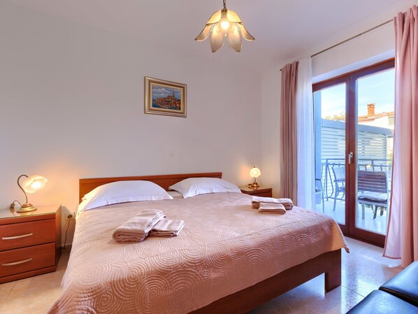 A neatly made bed with a pink quilt, two bedside lamps, a framed picture on the wall, and a balcony with a chair and table. - Apartment Alba by Interhome (Rovinj)