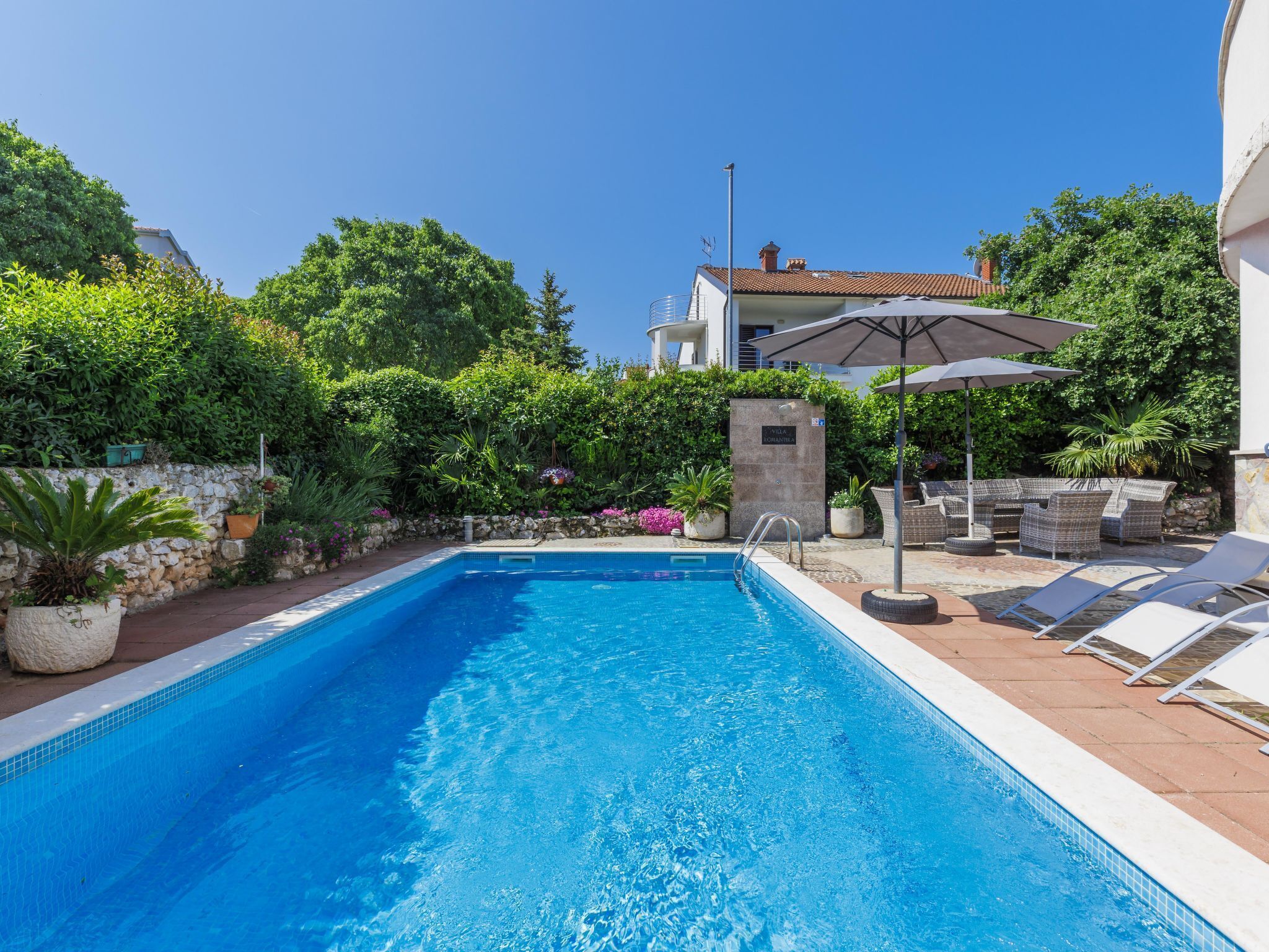 Piscina con muro de piedra y plantas en macetas. Hay sillas de descanso y una mesa con sombrillas.