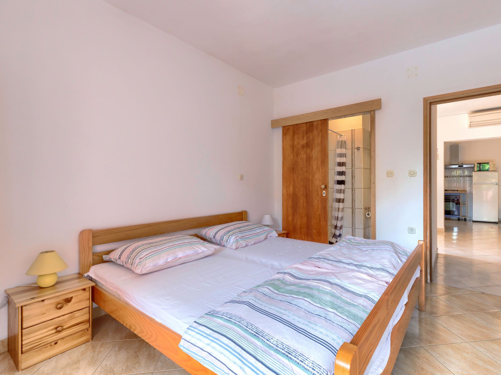 A bedroom with a wooden bed, a nightstand, a lamp, and a view of the bathroom and kitchen through an open door.