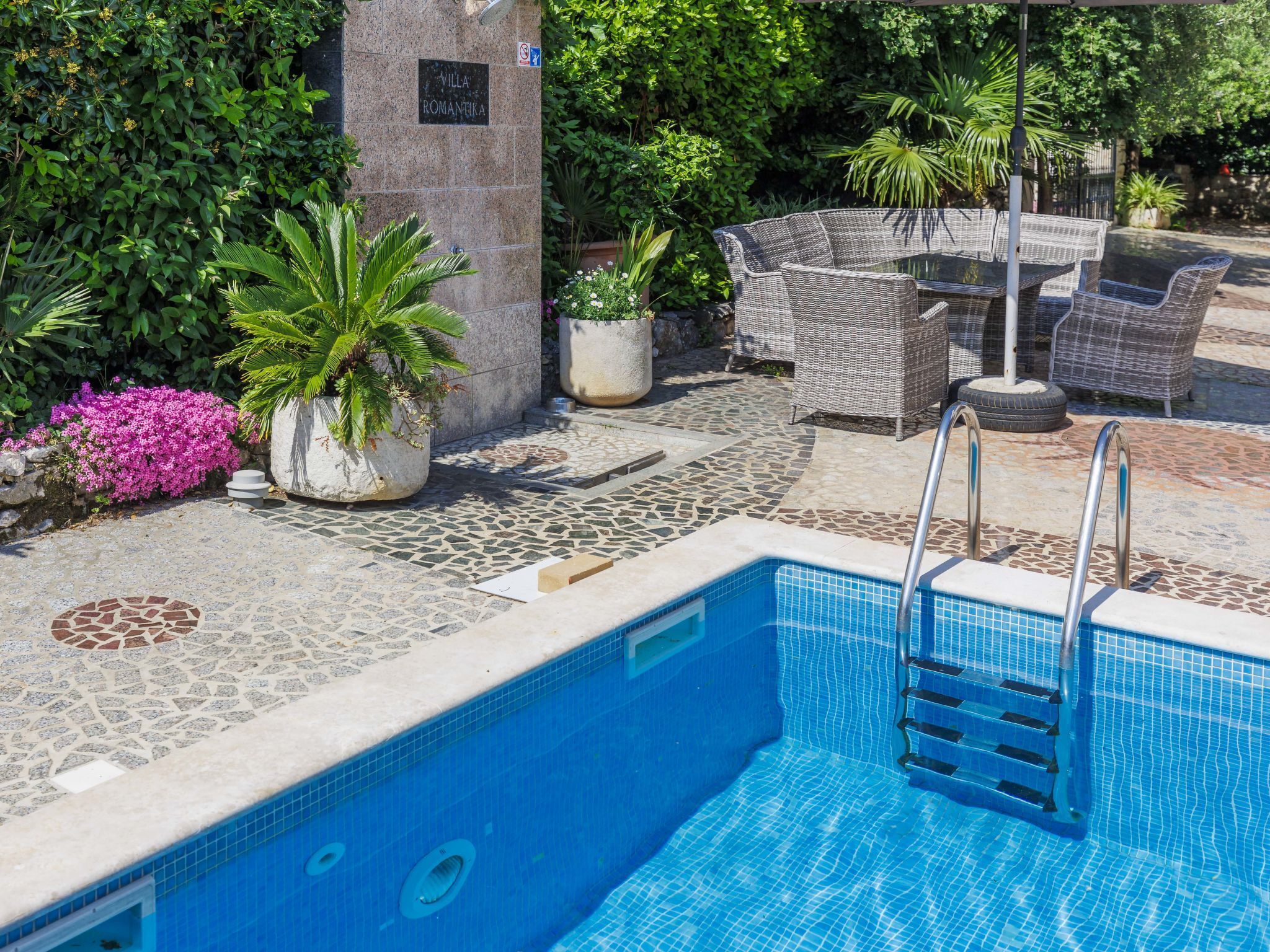 Una piscina con suelo de piedra, rodeada de exuberante vegetación y mobiliario de exterior.