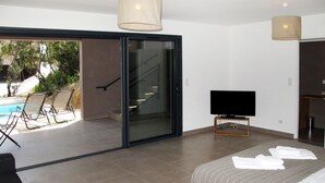 A modern bedroom with a large bed, a TV, and a sliding glass door leading to a balcony with chairs and a pool.