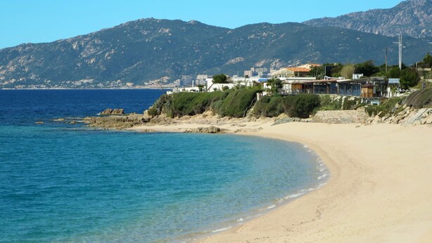 A beach with clear blue water, sandy shore, and buildings in the background.