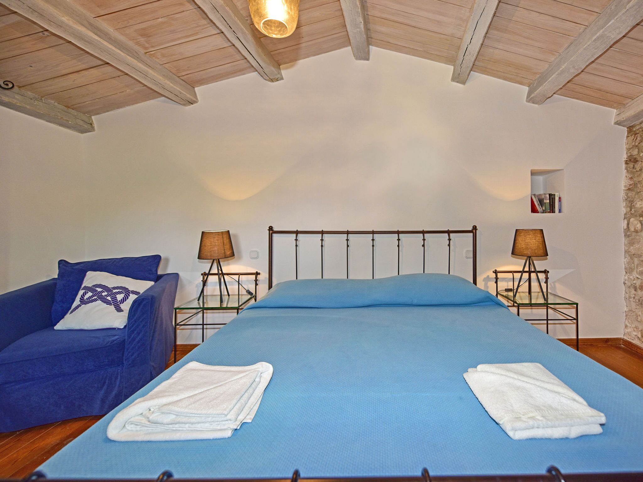Une chambre à coucher avec un lit, une chaise bleue, deux tables de chevet avec des lampes et une petite étagère avec des livres.