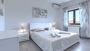 Un dormitorio con una cama grande, dos mesitas de noche, un aire acondicionado de pared y una ventana con cortinas.
