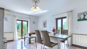 Un comedor con mesa, sillas y una araña. Se ve una ventana con cortinas, un radiador y un reloj en la pared.