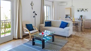 Un salon avec une table basse en verre, un canapé avec des coussins bleus, un coin repas avec une table et des chaises, et un balcon avec des rideaux.