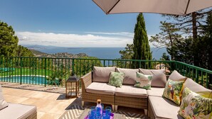 A balcony with a wicker sofa, a table with drinks, and a view of the sea and mountains. - Ponderosa by Interhome (Théoule sur mer)