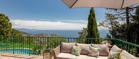 A balcony with a wicker sofa, a table with drinks, and a view of the sea and mountains.