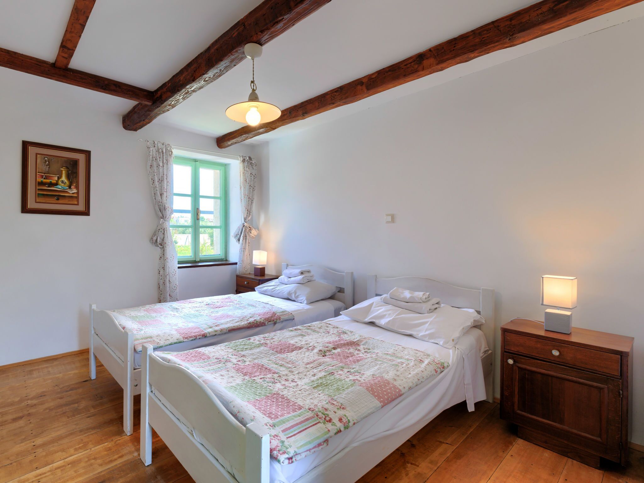 Une chambre avec deux lits simples, des poutres en bois au plafond, une fenêtre avec des rideaux, une table de chevet avec une lampe et un tableau encadré au mur.