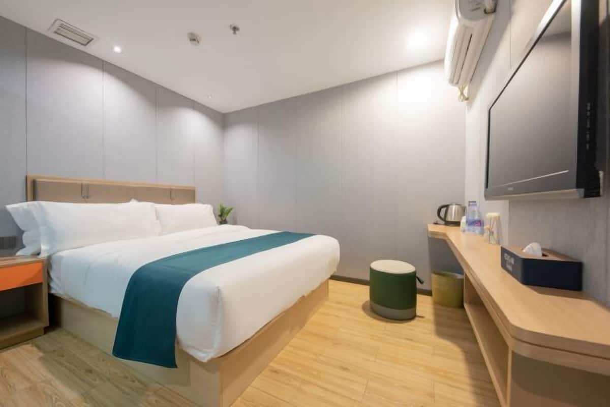 A modern hotel room with a bed, a small bedside table, a green waste bin, a wooden nightstand, and a flat-screen TV mounted on the wall.