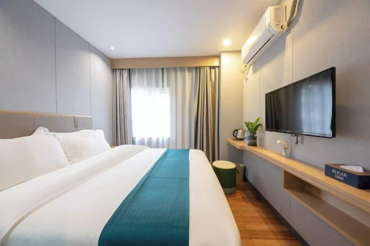 A hotel room with a large bed, a flat-screen TV mounted on the wall, a small table with a plant, and a window with curtains.